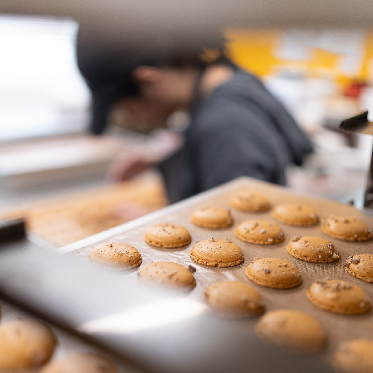 Baked macarons on a tray with a blurred background of a person and kitchen equipment.