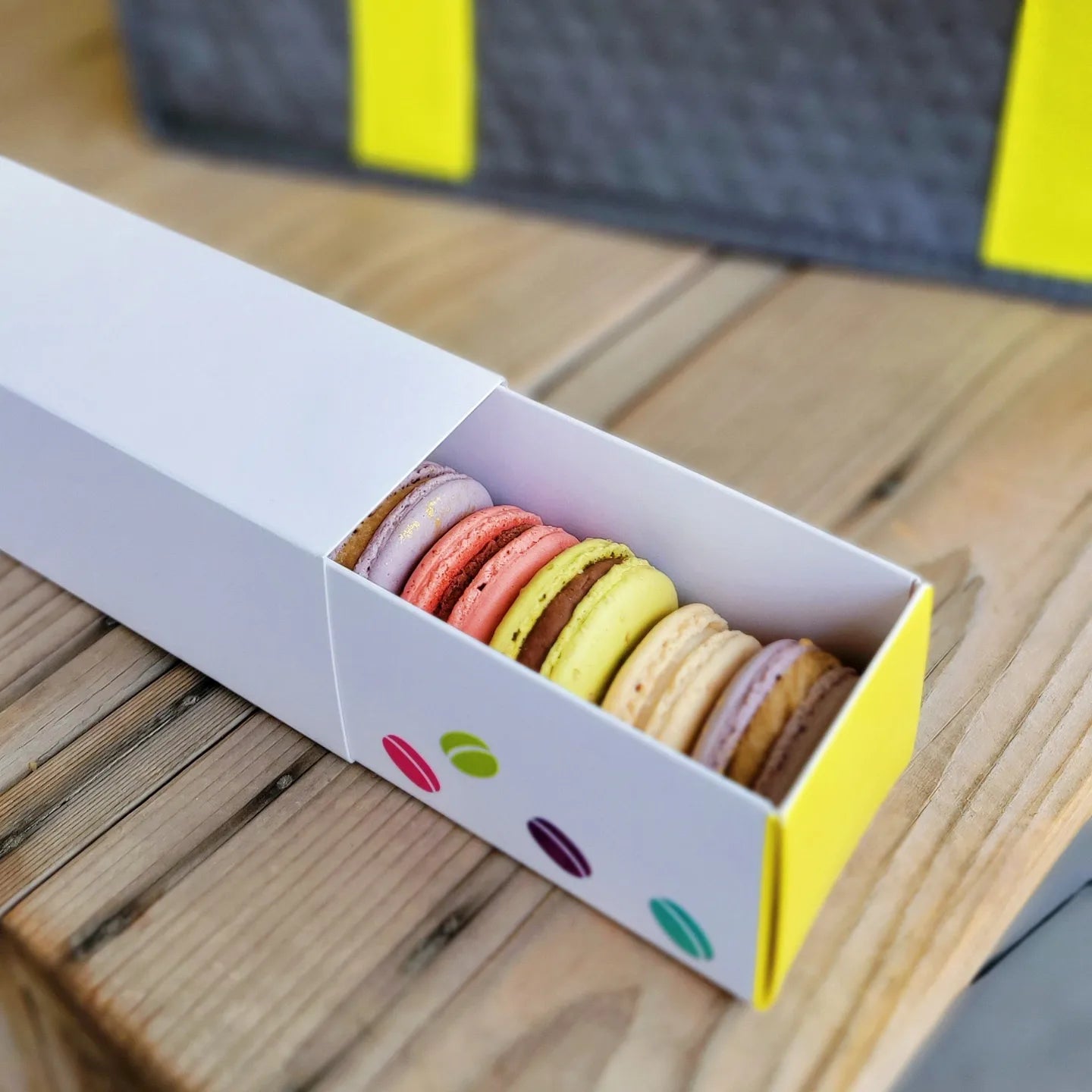 A white rectangular box with a yellow side panel and colorful macarons visible inside, placed on a wooden surface.