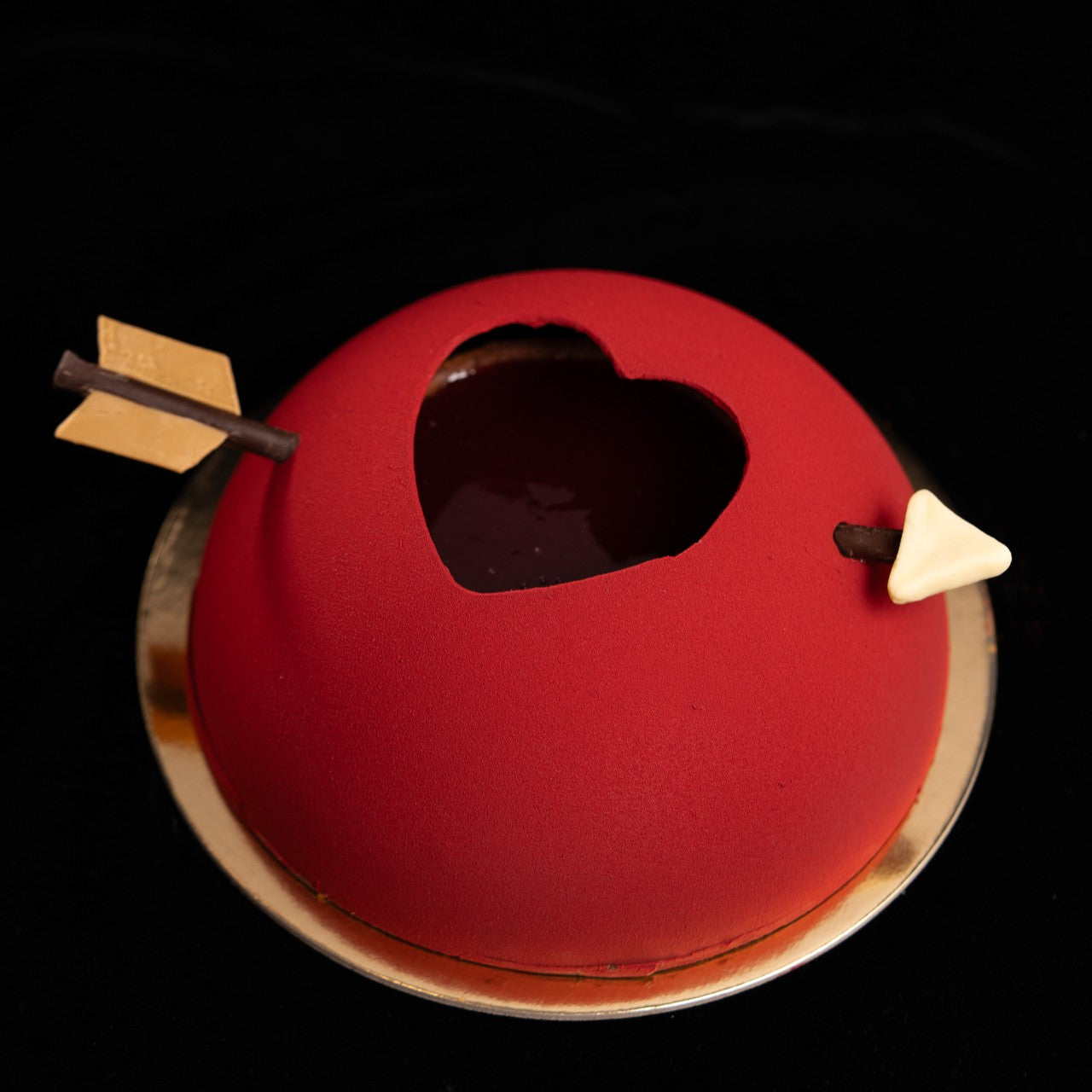 The chocolate semi-sphere features an open heart cut-out revealing its chocolate and raspberry cake core, pierced by a chocolate arrow straight through the heart.