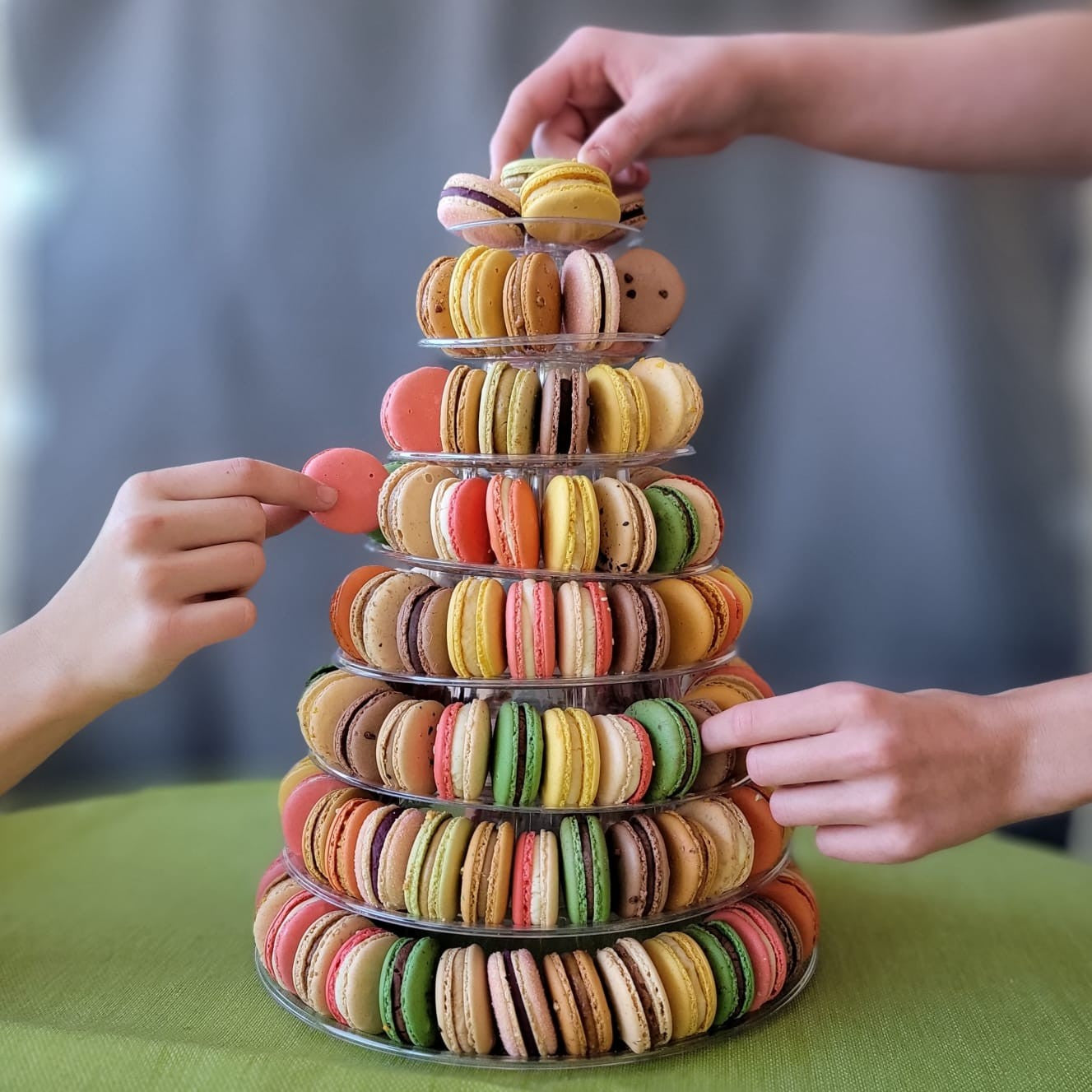 Colorful macaron tower with hands reaching to touch it on a green surface.