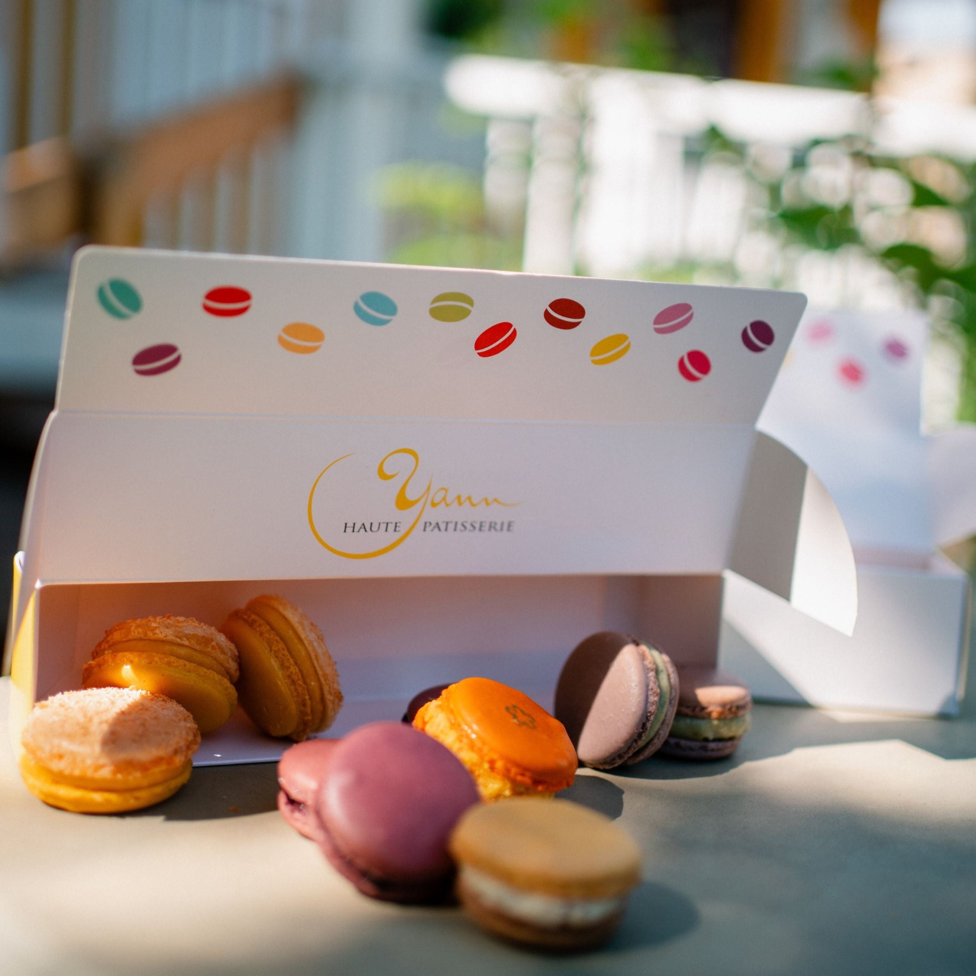 An open white pastry box with the brand name 'Vana Haute Patisserie' printed in gold, filled with assorted colorful macarons. More macarons are placed in front on a light surface, with a softly blurred outdoor patio background.