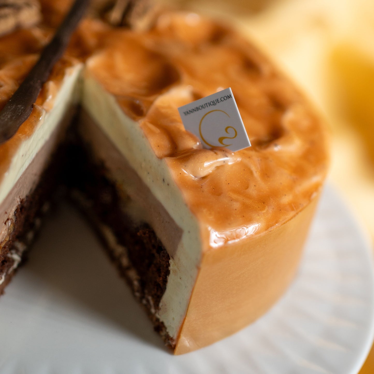 Close-up of a layered dessert with a slice missing, featuring a Yann Haute Patisserie brand tag.