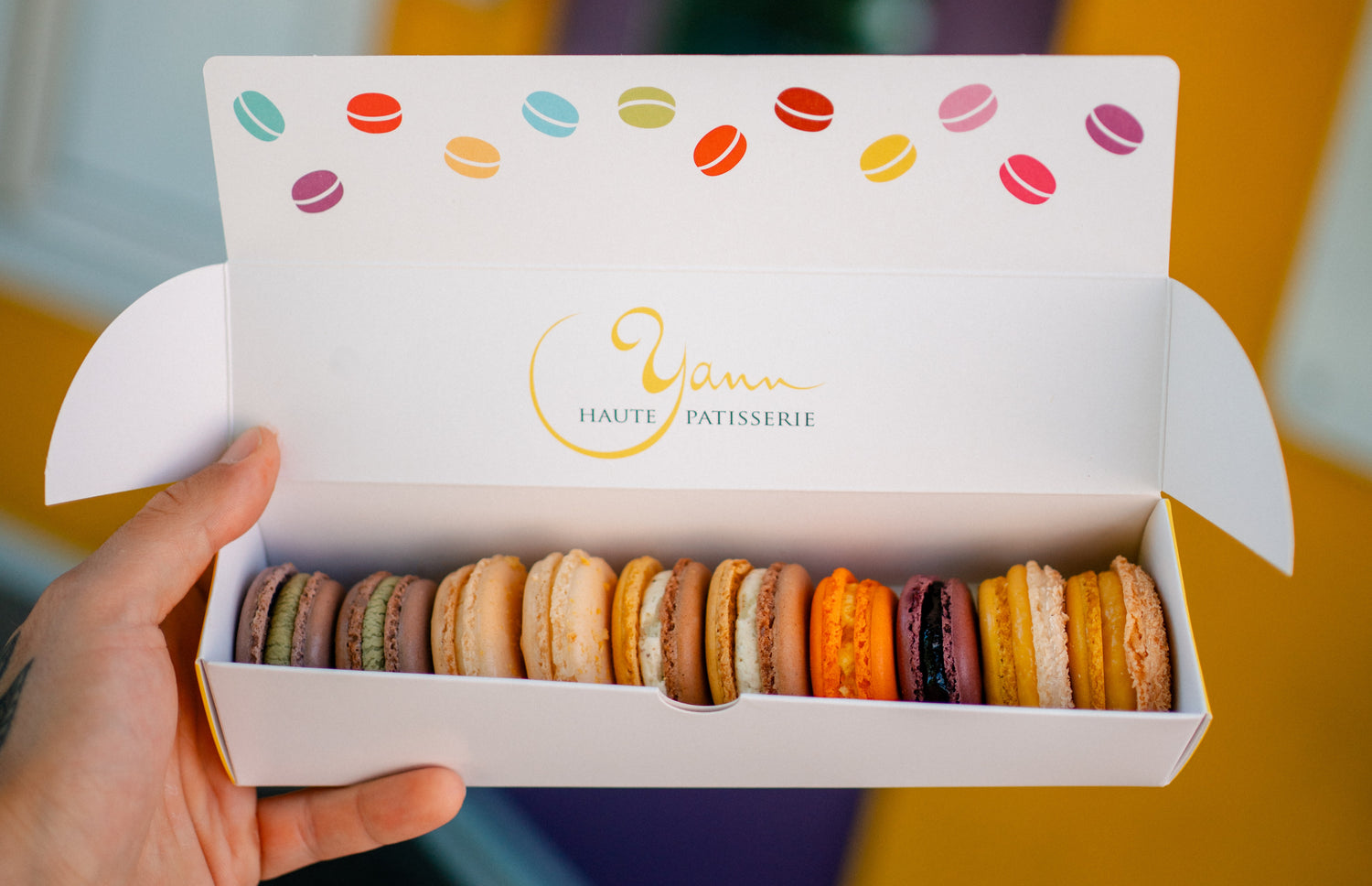 Box of colorful macarons held by a hand with 'Yann Haute Patisserie' branding.