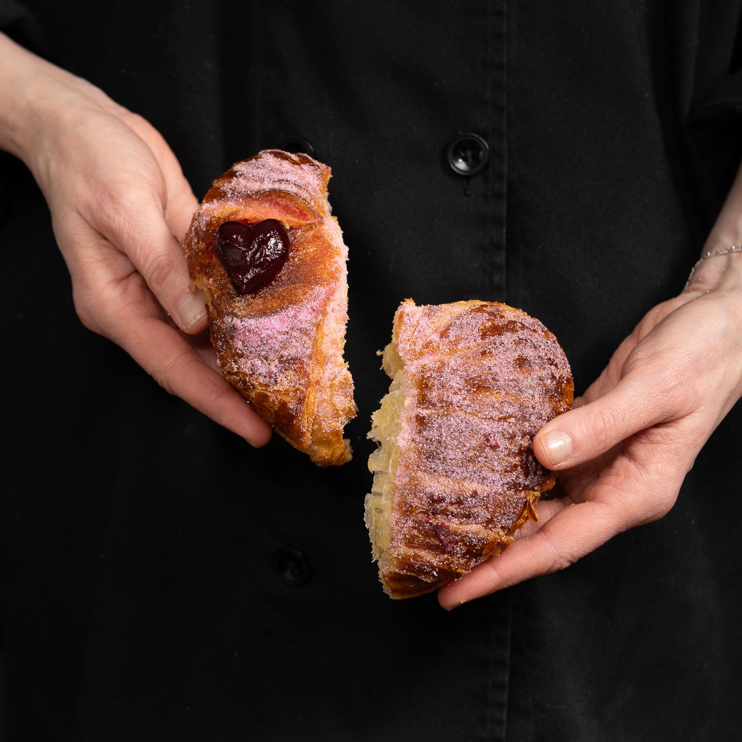 Two hands splitting a heart shaped croissant. both halves are filled with red berry compote!