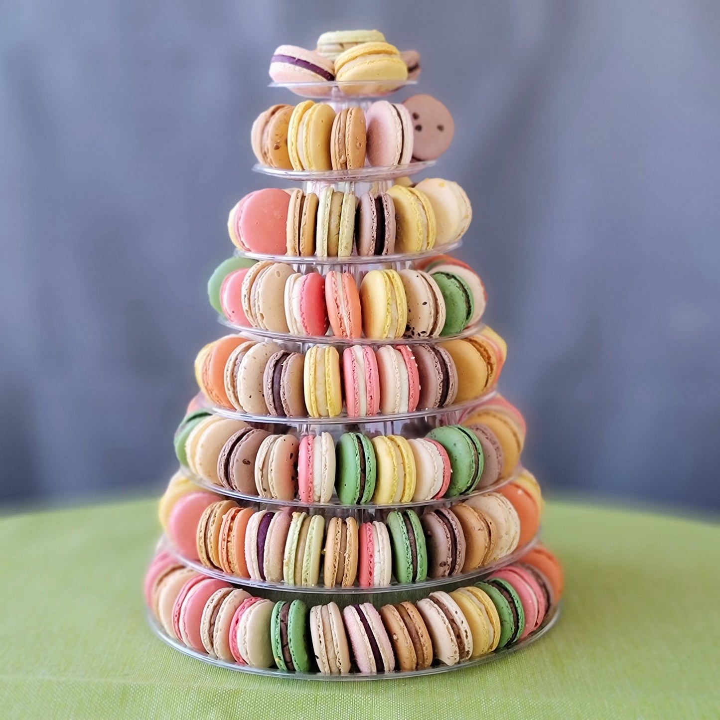 A pyramid-shaped tower of colorful macarons displayed on a clear stand against a grey background with 8 tiers!