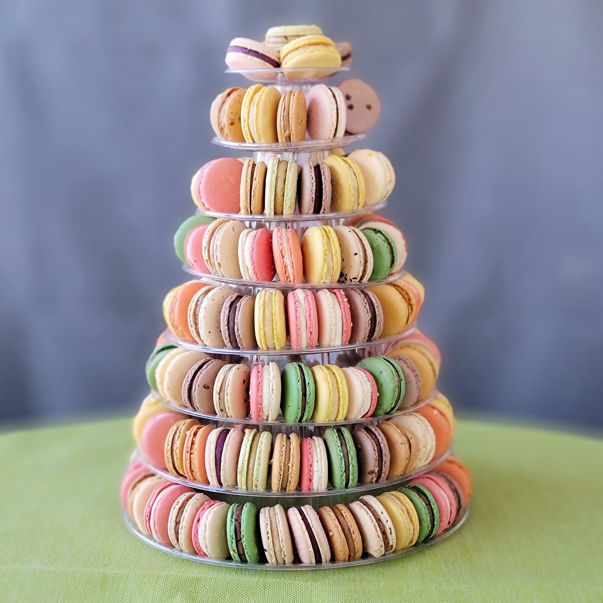 A pyramid-shaped tower of colorful macarons displayed on a clear stand against a grey background with 8 tiers!