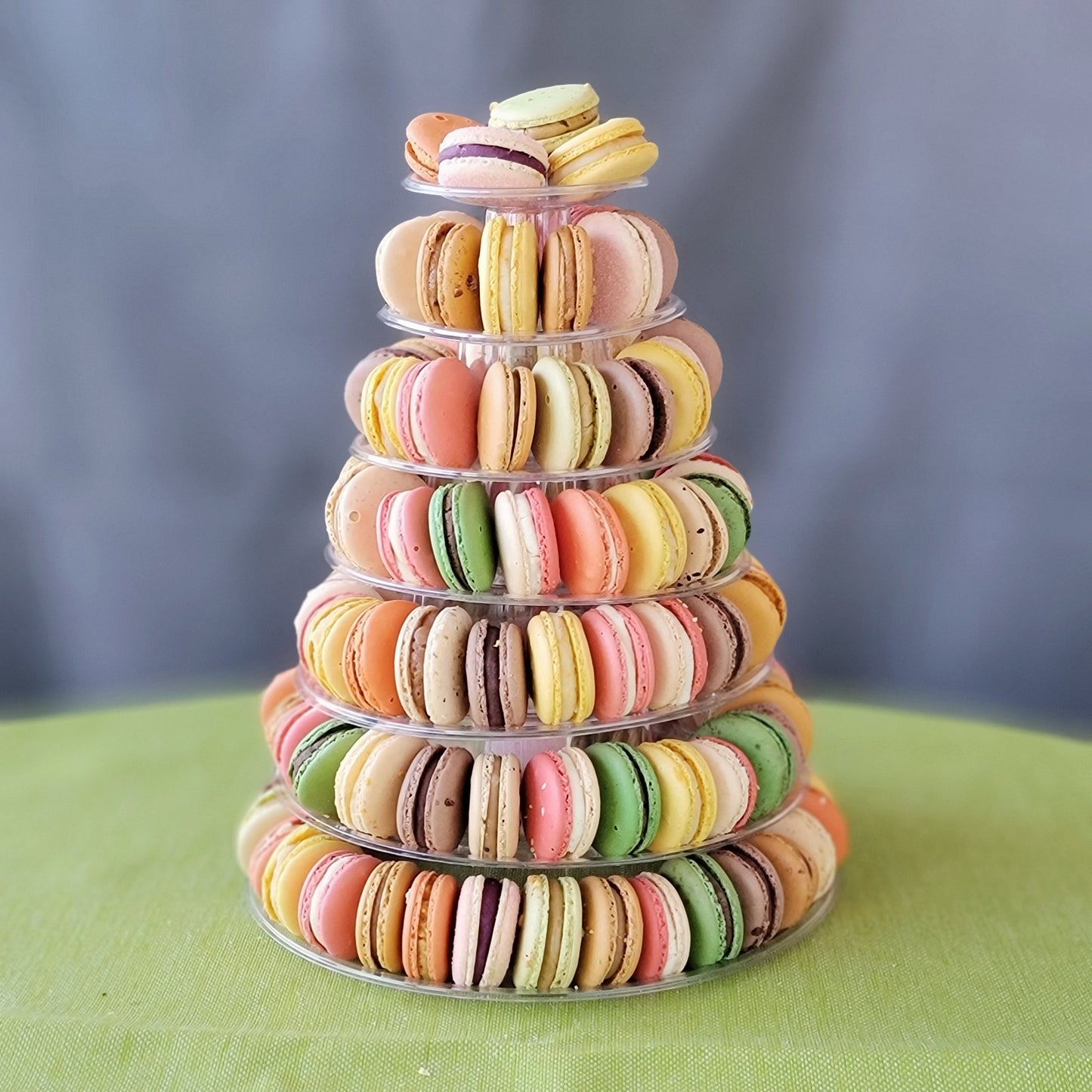 A pyramid-shaped tower of colorful macarons displayed on a clear stand against a grey background with 7 tiers!